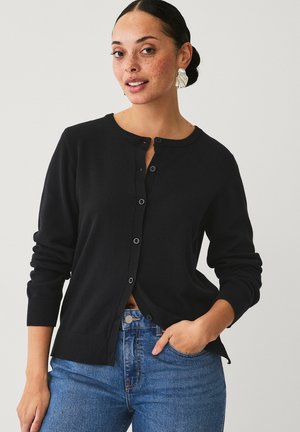 Gilet tricoté noir avec col rond, manches longues et fermeture à boutons. Présente des poignets et un ourlet côtelés. Associé à un pantalon en denim bleu.