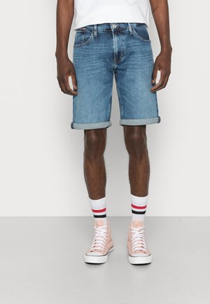 Mężczyzna w granatowych szortach dżinsowych z podwiniętymi nogawkami, białych skarpetach w czerwone i czarne paski oraz jasnoróżowych sneakersach za kostkę, stojący na szarym podłożu.