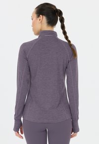 Kvinna med långt flätat hår som bär en åtsittande lila idrottströja med lång ärm och matchande leggings, sedd bakifrån.