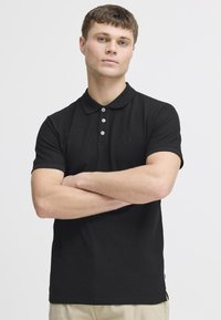 Sort polo-shirt med korte ærmer og ribbet krave. Har tre hvide knapper og et diskret logo på brystet. Lavet af bomulds materiale.