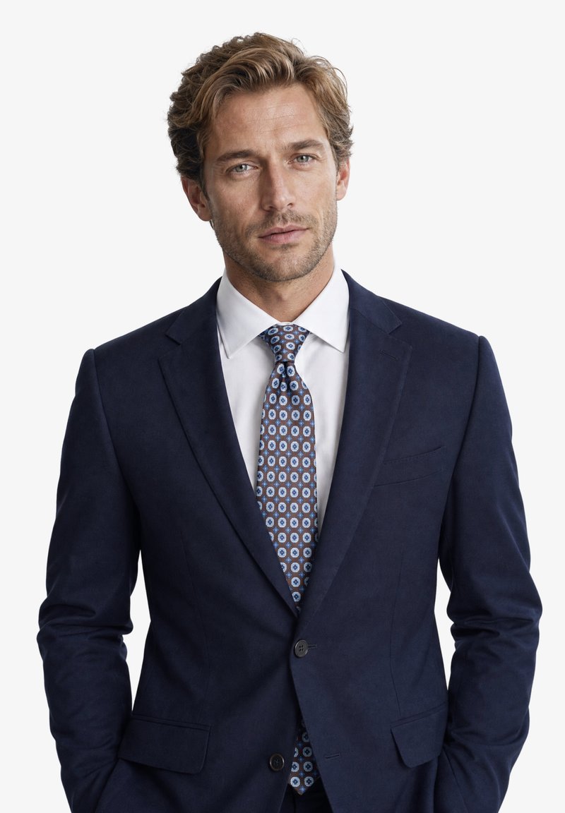 Homme blond portant une veste de costume bleu marine, une chemise blanche et une cravate à motifs bleu et marron, debout les mains dans les poches sur fond blanc.