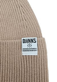 Beżowa czapka dzianinowa z prążkami, z białą metką z czarnym tekstem, na której widnieje napis "DJiNNs, 2001, Dreamed | Designed & Engineered in MH-City."