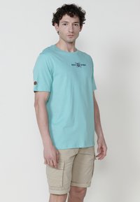 Himmelblaues Baumwoll-T-Shirt mit kurzen Ärmeln und rundem Ausschnitt, das auf der Vorderseite ein bedrucktes Logo und einen braunen Stoffakzent am Ärmel aufweist.