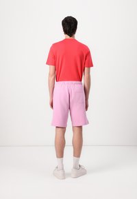 Röd t-shirt och rosa shorts med dragsko i midjan, med en bakficka. Modellen står med en neutral hållning mot en enkel bakgrund.