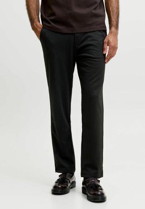 Mann trägt schwarze gerade geschnittene Hose, schwarze Tassel-Loafers und ein dunkelbraunes T-Shirt mit den Händen in und neben den Taschen.
