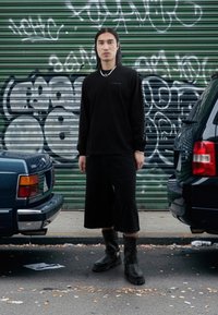 Černá košile s dlouhým rukávem s nápisy na rukávech, černé široké šortky a černé kozačky ke kolenům, stojící mezi dvěma auty před zdí s graffiti.