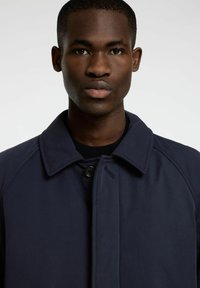 Mornarsko moder plašč s točkovnim ovratnikom, gumbom za zapiranje in raglan rokavi. Gladko, matirano blago z diskretno teksturo in brez vidnih vzorcev.