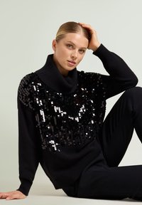 Suéter negro con cuello alto tipo cuello vuelto y frente decorado con lentejuelas. El diseño incluye un corte holgado y mangas largas.