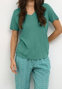 Tyrkysové tričko s výstřihem do V z měkkého materiálu, v kombinaci s volnými kalhotami s vzorem v geometrickém designu v tyrkysových a krémových barvách.