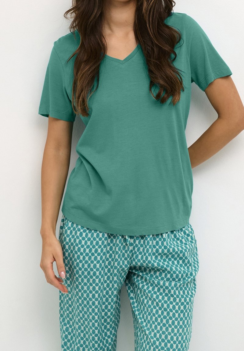 Tyrkysové tričko s výstřihem do V z měkkého materiálu, v kombinaci s volnými kalhotami s vzorem v geometrickém designu v tyrkysových a krémových barvách.