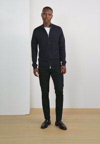 Svart zip-up tröja av stickat tyg, i kombination med slim-fit svarta byxor och svarta läderskor. Enkel design, inga synliga mönster.