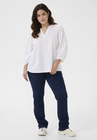 Weiße, leichte Bluse mit vertikaler Rippstruktur und V-Ausschnitt. Kombiniert mit dunkelblauen Jeans und beigen Sneakers.