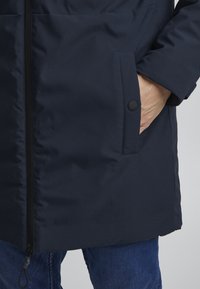 Marineblå, isolert jakke med glidelås foran, sidelomme med knappelukking, og en glatt tekstur, delvis synlig over denimjeans.