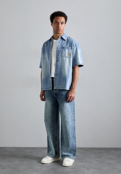 Denim skjorte med korte ermer i lyseblå gradient, frontlomme og broderte detaljer. Kombinert med vide blå jeans og hvite joggesko.