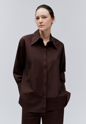 Giovane donna con camicia marrone scuro abbottonata e pantaloni coordinati, mani in tasca, espressione neutra, sfondo grigio chiaro uniforme.