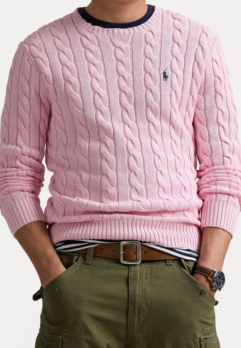 Personne portant un pull en maille torsadée rose clair, un pantalon vert olive avec les mains dans les poches, une ceinture en cuir marron et une montre-bracelet sombre.