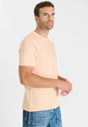 Homme portant une chemise beige à manches courtes et un jean bleu, debout les mains jointes, regardant légèrement sur le côté sur un fond blanc uni.