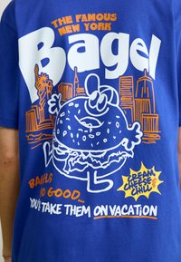 Blauwe katoenen t-shirt met witte en oranje grafische tekst, een cartoonbagel en een stadslandschap. De tekst luidt "De Beroemde New York Bagel."