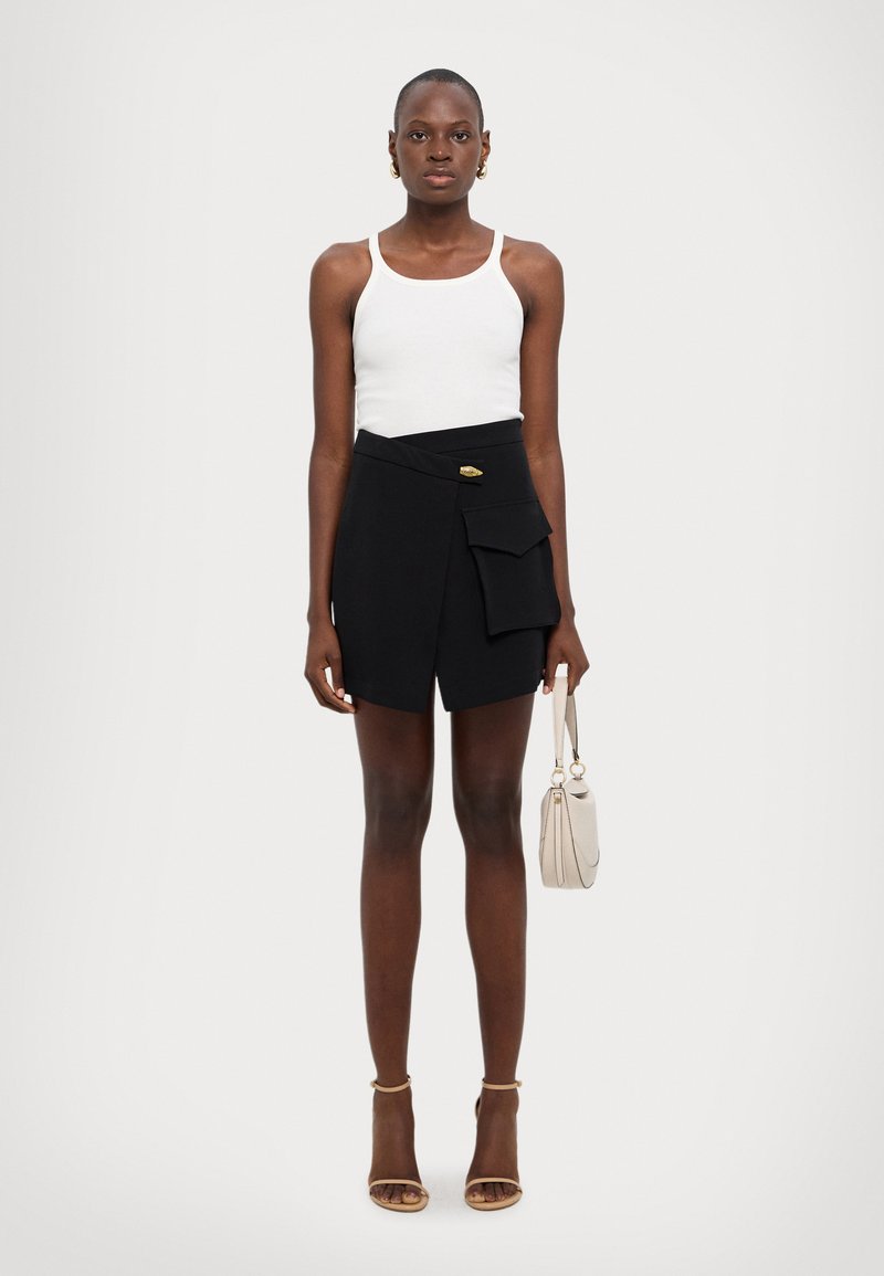 Czarna spódnica w owijanym stylu z boczną kieszenią w zestawie z białym topem na ramiączkach. Modelka trzyma małą beżową torebkę i ma na sobie beżowe sandały na paski.