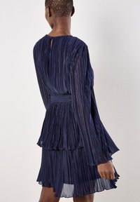 Granatowa plisowana sukienka z długimi, rozszerzającymi się rękawami, warstwowym dołem i otworem w kształcie klucza z tyłu; lekka, teksturowana tkanina o połysku.
