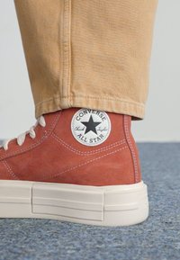 Zapatilla alta de Converse en ante rojo con suela de goma blanca y parche de logo en forma de estrella negra en el tobillo. Costuras detalladas visibles.