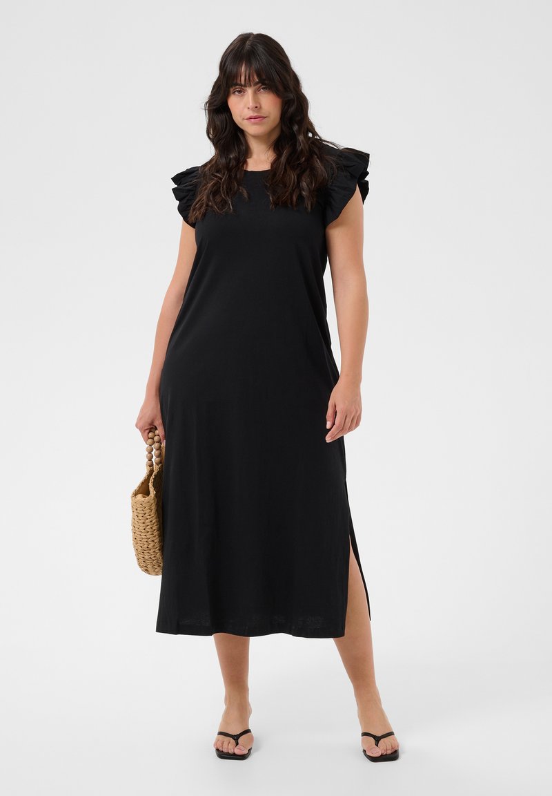 Woman wearing a sleeveless black midi dress with ruffled shoulders, holding a woven handbag, and wearing black sandals.