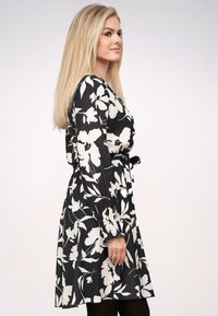Vestido negro con estampado floral blanco, de manga larga y cintura ceñida. Presenta puños texturizados con detalles. Se lleva con medias negras.
