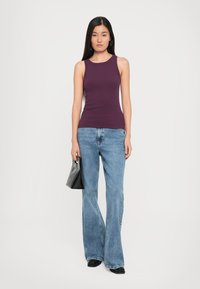 Paarse mouwloze top met subtiele reliëflogo, gecombineerd met lichtblauwe uitlopende jeans. Het model houdt een zwarte handtas vast.