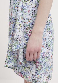 Bras droit et main reposant à côté d'une robe à motif floral multicolore avec des détails bleus, verts, roses et orange sur tissu blanc.