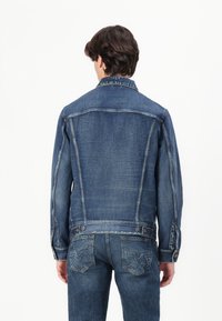 Denim jacket in dark blue with classic cut, button closure, and two back pockets. Features stitching details and slightly faded texture.