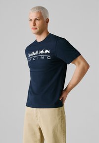 Junger Mann mit kurzen blonden Haaren, der ein dunkelblaues Red Bull Racing T-Shirt und beige Hosen trägt, mit einer Hand hinter dem Rücken stehend.