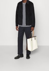 Giacca in velluto nero, camicia a righe, pantaloni blu navy, stivaletti neri alla caviglia e una borsa a tracolla bicolore (crema e marrone) con marchio di moda.