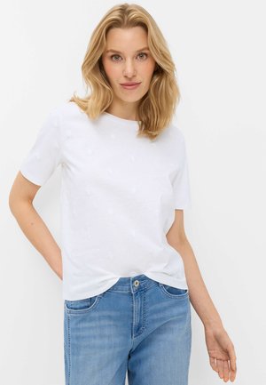 Blonde vrouw met een wit shirt met korte mouwen met subtiele geborduurde bloemen en lichtblauwe spijkerbroek, staand tegen een effen witte achtergrond.