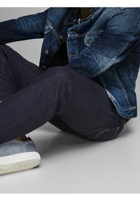 Jeansjacka med blå tvätt, guldfärgade knappar, kombinerad med mörka jeans och grå sneakers med vita sulor. Sittande pose mot en neutral bakgrund.