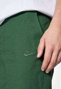 Green Nike pants feature a smooth texture, slim fit, side pockets, and an embroidered logo in the same shade. Hand resting on the fabric.