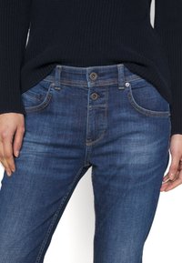 Mörkblå denimjeans med smal passform, två knappar och kontrasterande orange sömnad. Framfickorna synliga med lätt textur.