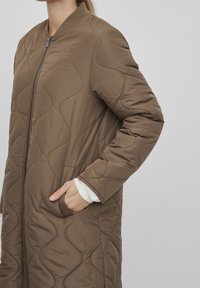 Brun vadderad lång jacka med ribbat krage, sidofickor och dragkedja. Har en slät textur och vågmönster på tyget.