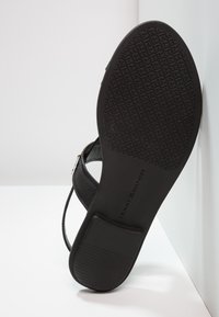 Sandale noire montrant une semelle texturée et un talon carré, reposant contre une surface blanche.