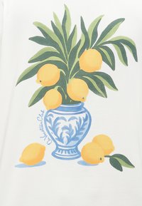 Modrý a biely keramický váza držiaci zelené listy a sedem žltých citrónov, s tromi citrónmi umiestnenými vedľa vázy na bielom pozadí.