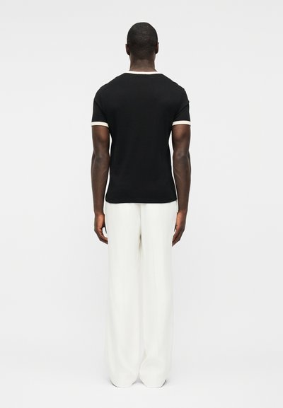 Άνδρας που κοιτάει προς τα πίσω φορώντας εφαρμοστή μαύρη μπλούζα με λευκή επένδυση στα μανίκια και στον γιακά, και άσπρο φαρδύ παντελόνι.