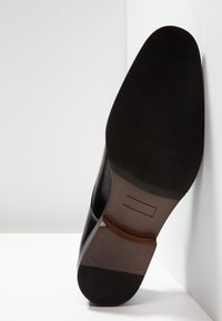 Chaussure en cuir noir avec une tige lisse et un détail de talon marron texturé. Dispose d'une semelle plate noire et d'une couture discrète.