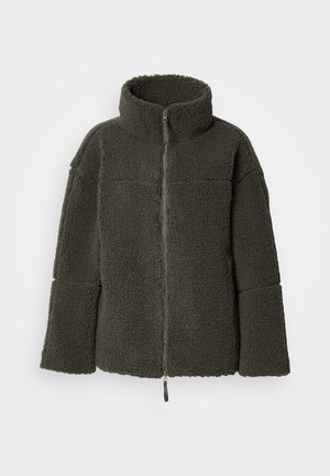 Flis jakna u tamnozelenoj boji, s uspravnim ovratnikom, punim prednjim patentnim zatvaračem i prevelikim dugim rukavima s teksturiranom površinom.