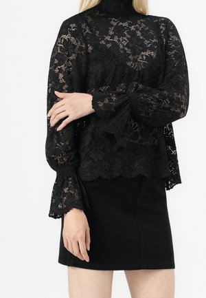 Femme portant un haut à manches longues en dentelle noire avec des motifs floraux et une jupe noire cintrée sur un fond neutre.