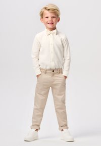Chemise à manches longues crème clair, pantalon en coton beige et baskets blanches. La chemise est ornée d'un nœud papillon et les pantalons sont retroussés aux chevilles.