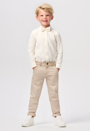 Chemise à manches longues crème clair, pantalon en coton beige et baskets blanches. La chemise est ornée d'un nœud papillon et les pantalons sont retroussés aux chevilles.