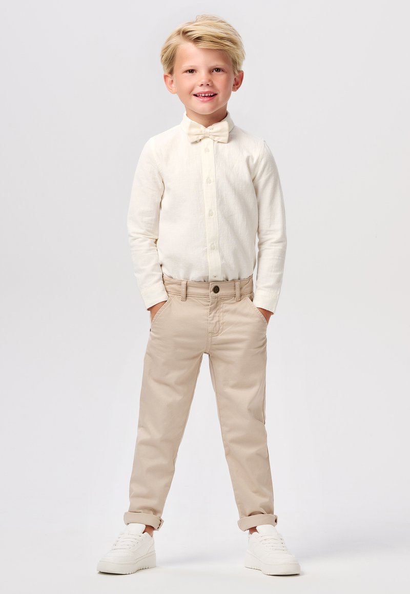 Chemise à manches longues crème clair, pantalon en coton beige et baskets blanches. La chemise est ornée d'un nœud papillon et les pantalons sont retroussés aux chevilles.