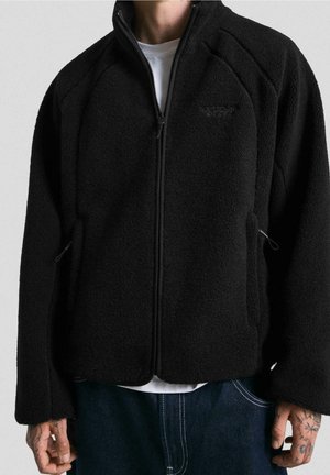 Person trägt eine schwarze Fleecejacke mit Reißverschluss über einem weißen Hemd, mit tätowierten Händen und sichtbaren blauen Jeans.