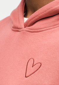 Ροζ φούτερ με απαλή υφή, διακοσμημένο με μια καφέ καρδιά κεντημένη στο στήθος και διπλή φόδρα στην κουκούλα.