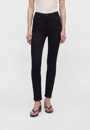 Kvinde iført sort skinny jeans med høj talje, kombineret med brune kilehæls-sandaler og en beige ærmeløs top, stående mod en ensfarvet baggrund.