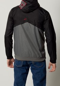 Svart och grå jacka med huva, med ett unikt designelement på baksidan, elastiska manschetter och en grå midja.